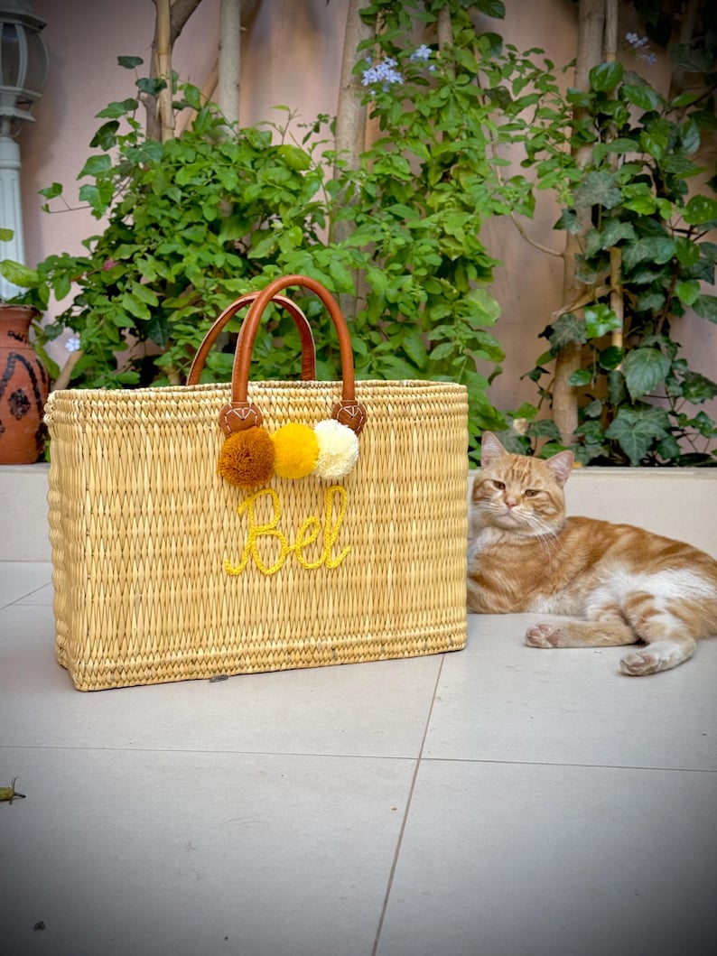 May include: A rectangular woven straw tote bag with brown leather handles and three pom-poms in brown, yellow, and white. The word "Bel" is embroidered in yellow on the front. An orange tabby cat rests nearby.