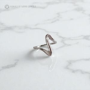 Puede incluir: Un anillo plateado con un diseño abstracto único. El anillo presenta una forma fluida y abierta con una parte superior puntiaguda y una base curva. El anillo está sobre una superficie de mármol blanco. La marca "SORELI STUDIO JEWELS" está en la esquina superior izquierda.