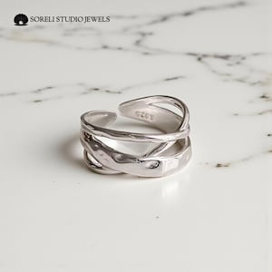Op de afbeelding: Een zilverkleurige ring met een getextureerd, overlappend ontwerp. De ring heeft een gehamerd uiterlijk en heeft de tekst "S925" aan de binnenkant. De merknaam "Soreli Studio Jewels" is zichtbaar in de linkerbovenhoek.