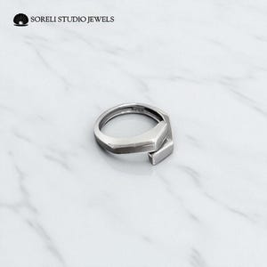 May include: A silver ring with a geometric design. The ring features a rectangular bar that overlaps a section of the band. The words "SORELI STUDIO JEWELS" are printed in black.