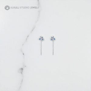 Puede incluir: Un par de pendientes de plata. Cada pendiente presenta un grupo de piedras preciosas azul claro y una larga y fina cadena de plata. Los pendientes se exhiben sobre una superficie de mármol blanco. El texto "SOBELI STUDIO JEWELS" está en la esquina superior izquierda.