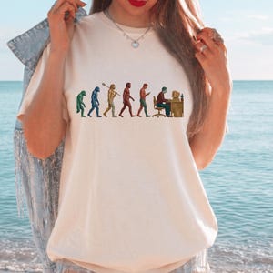 Pode incluir: Uma t-shirt de manga curta na cor creme com uma impress&atilde;o gr&aacute;fica da evolu&ccedil;&atilde;o humana. O design mostra uma progress&atilde;o de um primata verde para um primata azul, um homin&iacute;deo amarelo com uma lan&ccedil;a, um homin&iacute;deo castanho, uma figura vermelha e uma pessoa numa secret&aacute;ria.