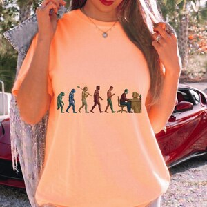 Pode incluir: Uma t-shirt cor de p&ecirc;ssego com uma impress&atilde;o gr&aacute;fica da evolu&ccedil;&atilde;o humana, come&ccedil;ando com um primata e progredindo atrav&eacute;s de v&aacute;rias fases at&eacute; uma pessoa sentada a uma secret&aacute;ria com um computador. A t-shirt tem um decote redondo.