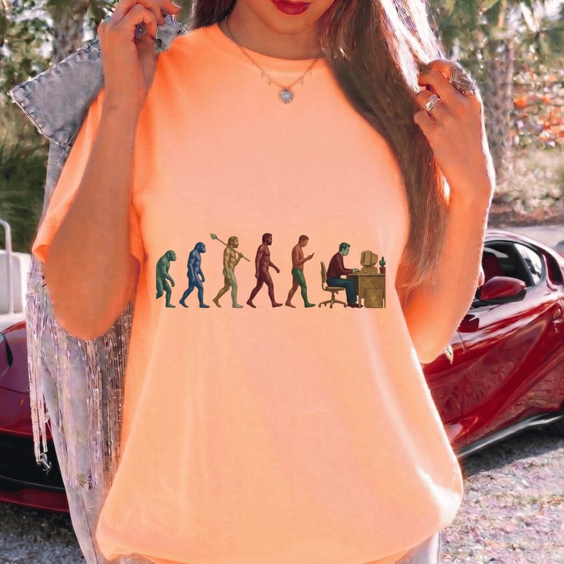 Pode incluir: Uma t-shirt cor de p&ecirc;ssego com uma impress&atilde;o gr&aacute;fica da evolu&ccedil;&atilde;o humana, come&ccedil;ando com um primata e progredindo atrav&eacute;s de v&aacute;rias fases at&eacute; uma pessoa sentada a uma secret&aacute;ria com um computador. A t-shirt tem um decote redondo.