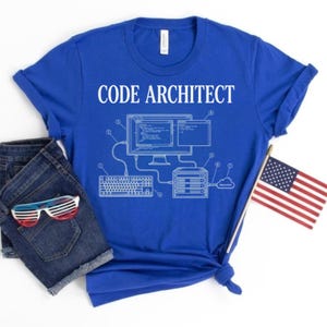 May include: A royal blue short-sleeve t-shirt featuring the white text CODE ARCHITECT above a white line-art graphic of a computer monitor, keyboard, and server tower. Dark blue denim shorts and a small American flag are positioned nearby.
