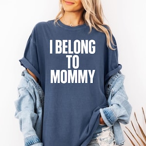 Peut inclure: Un t-shirt bleu marine avec le texte "I BELONG TO MOMMY" en lettres blanches. Le t-shirt a des manches retroussées et est porté avec une veste en jean. Le style général est décontracté et confortable.