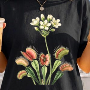 Op de afbeelding: Een zwart t-shirt met een botanische illustratie van een Venusvliegenvanger met groene en rode vallen, waarvan sommige insecten bevatten, en een tros witte bloemen aan de bovenkant. De afbeelding is gecentreerd op de borst van het kledingstuk.