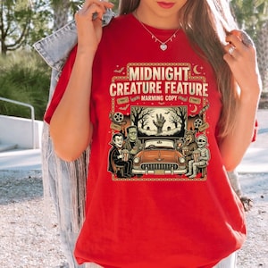 Pode incluir: Uma t-shirt vermelha com uma estampa gráfica intitulada MIDNIGHT CREATURE FEATURE MARMING COPY. O design retrata uma cena de cinema drive-in com um carro clássico, um vampiro, Frankenstein, um lobisomem e uma múmia a comer pipocas.