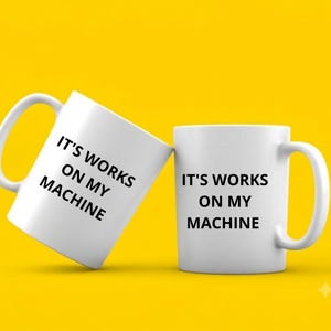 May include: Two white ceramic mugs featuring the black text IT'S WORKS ON MY MACHINE printed on the front. One mug is positioned upright while the other is tilted at an angle, both displaying the same phrase in a clear, sans-serif font.
