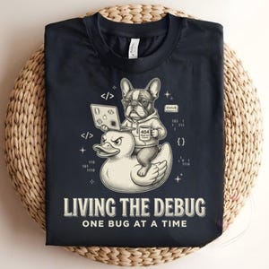 Op de afbeelding: Een donkerblauw T-shirt met een afbeelding van een Franse bulldog met een bril en hoodie, zittend op een badeendje terwijl deze een laptop gebruikt. Het ontwerp bevat codesymbolen, binaire getallen en de tekst "LIVING THE DEBUG ONE BUG AT A TIME".