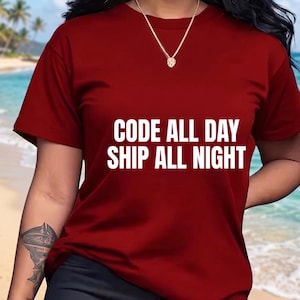 May include: A dark red short-sleeved t-shirt featuring the white text "CODE ALL DAY SHIP ALL NIGHT" printed across the chest. The shirt has a crew neckline and is worn with a gold chain necklace.
