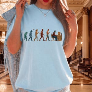 Pode incluir: Uma t-shirt azul claro com uma estampa gr&aacute;fica da evolu&ccedil;&atilde;o humana. O design mostra uma progress&atilde;o de um primata para um humano moderno sentado a uma secret&aacute;ria com um computador. A t-shirt tem um decote redondo e mangas curtas.