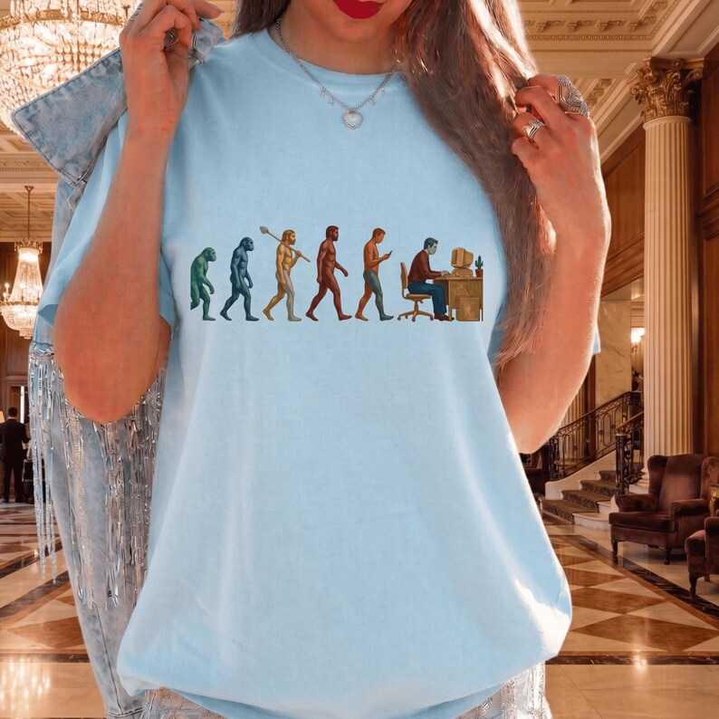 Pode incluir: Uma t-shirt azul claro com uma estampa gr&aacute;fica da evolu&ccedil;&atilde;o humana. O design mostra uma progress&atilde;o de um primata para um humano moderno sentado a uma secret&aacute;ria com um computador. A t-shirt tem um decote redondo e mangas curtas.