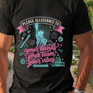 May include: A black t-shirt featuring a graphic print with the text PLEDGE ALLEGIANCE TO your hands your team your vibes. The design includes a Statue of Liberty, a guitar, a disco ball, a cat, fireworks, and the U.S. Capitol building in neon colors.
