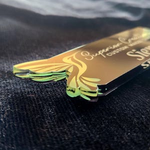 May include: A custom-made name tag with the text "Superior Custom Made Siena" in gold lettering. The tag has a unique shape with a decorative design in shades of yellow and green. The background is a dark, textured fabric.