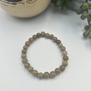 May include: A beaded bracelet featuring round, earthy-toned beads in shades of green and brown. The bracelet is strung on an elastic cord and is displayed on a white surface. The beads appear to be approximately 0.3 inches in diameter.