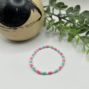 Puede incluir: Una pulsera de cuentas con cuentas rosas, celeste y blancas. La pulsera es circular y parece estar hecha de pequeñas cuentas uniformes ensartadas. La pulsera está sobre una superficie blanca.