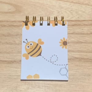 May include: A small, spiral-bound notepad with a bee design. The cover shows a cartoon bee with a smiling face, yellow wings, and black stripes. Additional graphics include a sunflower and honeycomb shapes. The notepad has gold-coloured spiral binding.