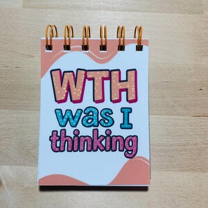 May include: A small spiral-bound notepad with the text "WTH was I thinking" in colourful bubble letters. The notepad has a peach and white design with orange spiral binding.