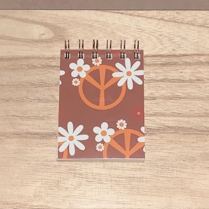 May include: A small spiral-bound notepad with a brown cover featuring white daisies and orange peace signs. The notepad is on a wooden surface.