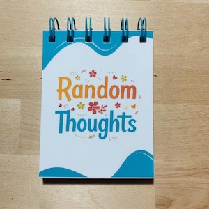May include: A small notepad with a teal cover and spiral binding. The cover features the words "Random Thoughts" in orange and teal, with floral and musical note accents. The notepad is on a wooden surface.