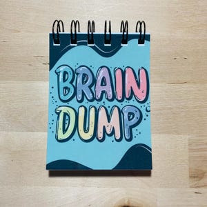 May include: A small, spiral-bound notepad featuring the words "BRAIN DUMP" in colourful bubble letters on a light blue background. The notepad has a black spiral binding and is set against a light wood grain surface.