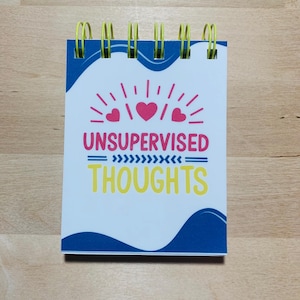 May include: A small notepad with yellow spiral binding. The cover is white with blue accents and the words "UNSUPERVISED THOUGHTS" in pink and yellow, with heart and sunburst graphics.