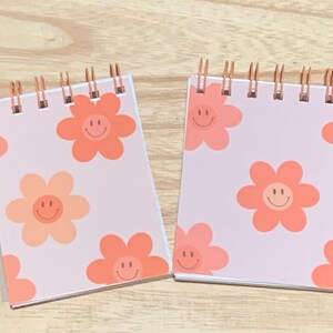 May include: Two small spiral-bound notepads with a light pink cover. Each notepad features a repeating pattern of orange flower designs with smiling faces. The spiral binding is copper-coloured.