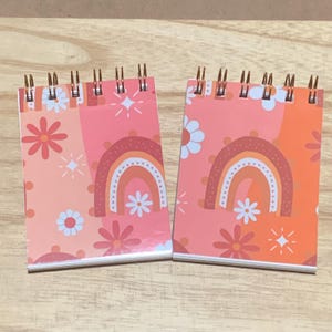 May include: Two small spiral-bound notepads with a retro design. Each notepad features a pink and orange colour scheme with a rainbow, flowers, and starbursts. The notepads are on a light brown wooden surface.
