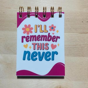 May include: A small spiral-bound notepad with a white cover featuring the phrase "I'll remember THIS never" in various colours. The design includes flowers, hearts, and decorative swirls in pink, orange, and blue.