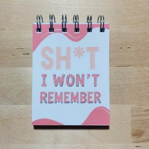 May include: A small, spiral-bound notepad with a white cover. The cover displays the text "SH*T I WON'T REMEMBER" in pink, with pink wavy accents and a sun graphic. The notepad is on a wooden surface.