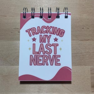 May include: A small, spiral-bound notepad with a white cover and pink accents. The cover features the text "TRACKING MY LAST NERVE" in a retro, illuminated font, with star and cross embellishments.