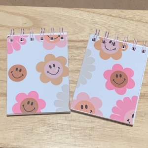 May include: Two small spiral-bound notebooks with a white background. Each notebook features a repeating pattern of smiling flower faces in shades of pink, peach, and brown. The notebooks have a retro, cheerful design.