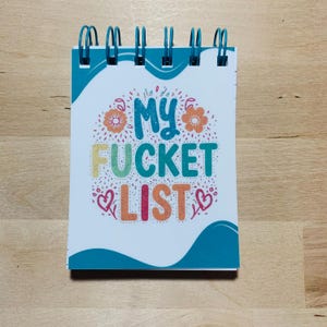 May include: A small notepad with a teal spiral binding. The cover features the words "My Fucket List" in colourful, playful lettering, surrounded by floral and heart illustrations. The notepad is on a wooden surface.
