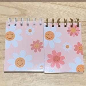May include: Two spiral-bound notebooks with pink covers featuring a daisy pattern with smiling faces. The daisies are white, pink, and orange. The notebooks are on a light wood surface.