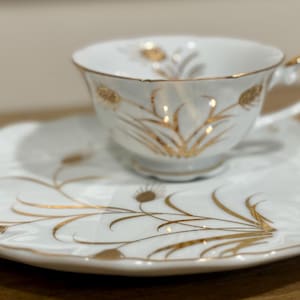 May include: A white teacup and saucer set with gold wheat stalk designs. The cup has a handle and sits on a matching saucer with a scalloped edge. The set is made of porcelain and is suitable for serving tea.