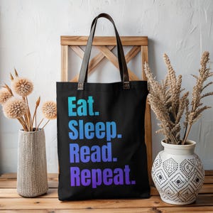 May include: Black tote bag with brown leather handles. The bag features the words "Eat. Sleep. Read. Repeat." in a gradient of blue and purple. The bag is displayed in front of a wooden background with decorative vases.
