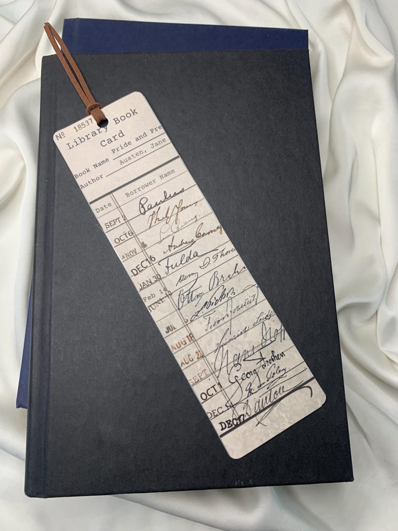 Op de afbeelding: Een vintage boekenlegger met een bruin leren koord ligt op een gesloten zwart boek. De boekenlegger is een bibliotheekkaart voor "Pride and Prejudice" van Jane Austen, met handgeschreven namen en data.