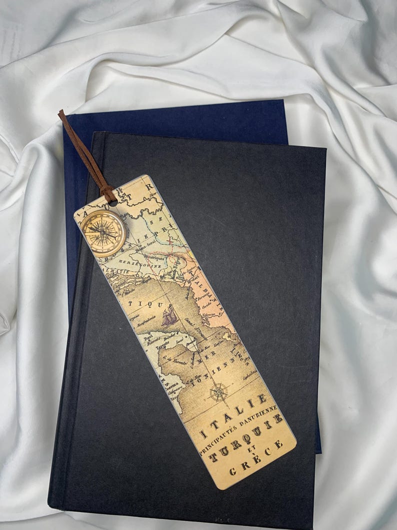 Op de afbeelding: Een vintage boekenlegger met een kaart en een kompas. Het heeft een bruin leren koord. De boekenlegger ligt op een donkerblauw en zwart boek, tegen een witte achtergrond.