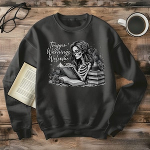 Könnte beinhalten: Dunkelgraues Sweatshirt mit einer Schwarz-Weiß-Illustration eines Skeletts, das ein Buch liest. Das Skelett trägt eine Brille und hat lange Haare. Der Text "Trigger Warnings Welcome" ist in weißer Schrift geschrieben. Ein Stapel Bücher und Rosen sind ebenfalls im Design enthalten.