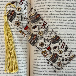 May include: Clear acrylic bookmark with gold tassel. The bookmark features illustrations of books, coffee cups, eyeglasses, and flowers. A decorative accessory for book lovers.