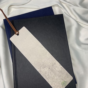 Op de afbeelding: Een rechthoekige bladwijzer met een lichtgrijze, getextureerde oppervlakte en een bruin leren koord. De bladwijzer ligt op een zwart boek met daaronder een marineblauw boek, alles op een witte stoffen achtergrond. Een klein groen cirkelvormig logo is zichtbaar.
