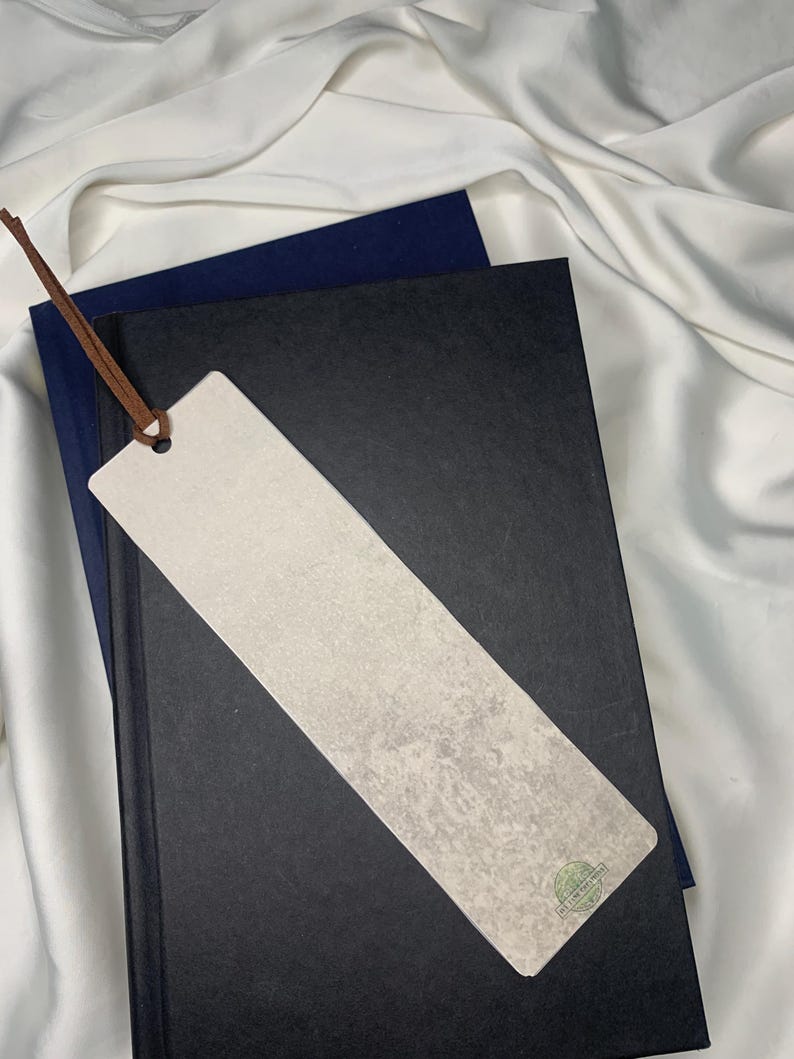 Op de afbeelding: Een rechthoekige bladwijzer met een lichtgrijze, getextureerde oppervlakte en een bruin leren koord. De bladwijzer ligt op een zwart boek met daaronder een marineblauw boek, alles op een witte stoffen achtergrond. Een klein groen cirkelvormig logo is zichtbaar.