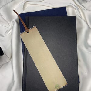 Op de afbeelding: Een beige bladwijzer met een bruin leren touwtje ligt op een zwart boek en een marineblauw boek. De bladwijzer heeft een decoratieve rand en een klein groen cirkelvormig ontwerp. De achtergrond is een witte, zijdeachtige stof.