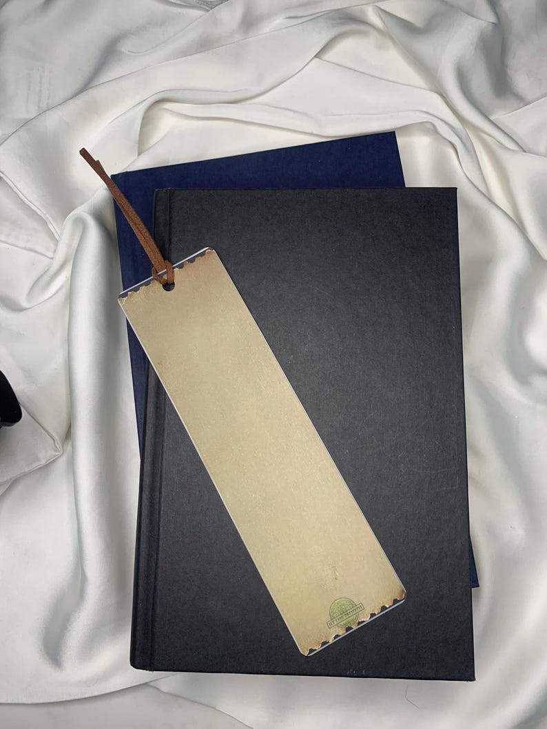 Op de afbeelding: Een beige bladwijzer met een bruin leren touwtje ligt op een zwart boek en een marineblauw boek. De bladwijzer heeft een decoratieve rand en een klein groen cirkelvormig ontwerp. De achtergrond is een witte, zijdeachtige stof.