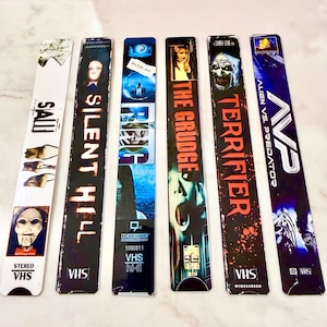 May include: Six vertical rectangular bookmarks featuring horror movie themes including Saw, Silent Hill, The Ring, The Grudge, Terrifier, and Alien vs. Predator. Each bookmark displays the film title, iconic imagery, and a VHS logo at the bottom.