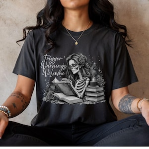 May include: Dark gray t-shirt featuring a skeleton reading a book, with the text "Trigger Warnings Welcome." The graphic includes roses, a candle, and a stack of books. The design is in black and white.