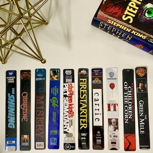 May include: A collection of vintage VHS tapes featuring Stephen King adaptations, including "The Shining," "Christine," and "Misery." The tapes are arranged with a Stephen King book and a gold starburst decoration.