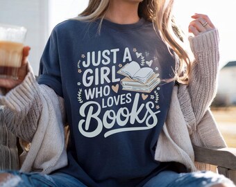 May include: Navy blue sweatshirt with the text "JUST A GIRL WHO LOVES BOOKS" in white. The design includes illustrations of books, hearts, and floral accents. The person is wearing blue jeans and a beige cardigan.