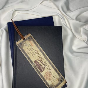 Op de afbeelding: Een vintage-stijl bladwijzer met de tekst "American Railway" en een treinillustratie. De bladwijzer heeft een leren koord en ligt op een zwart boek met een blauw boek erachter. De achtergrond is wit.